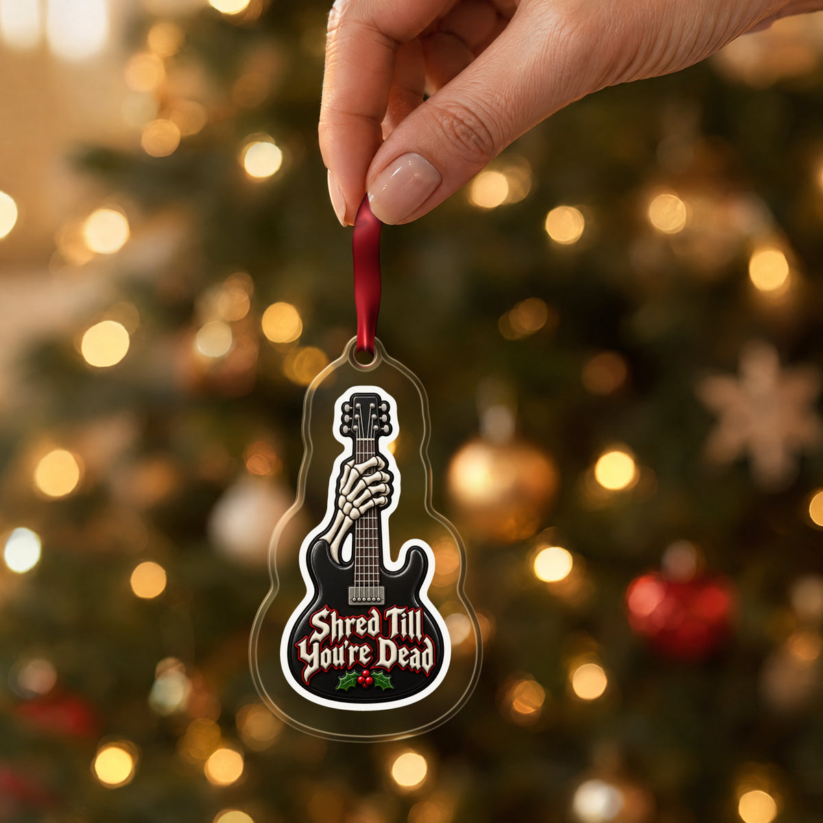 Shred Till You're Dead Skeleton Guitar clear acrylic Christmas ornament held by its red ribbon in a hand, shown at its real size