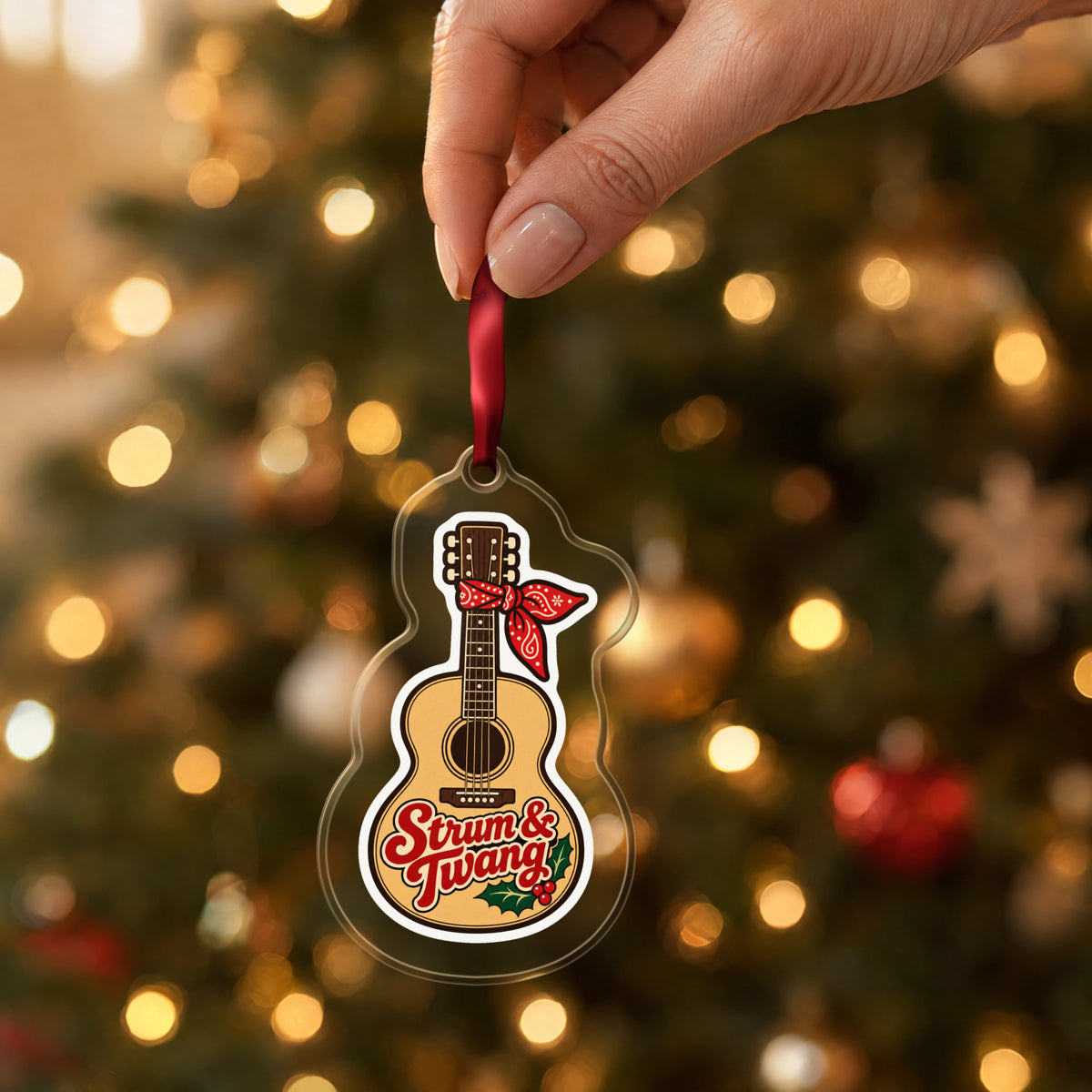 Strum & Twang Country Guitar clear acrylic Christmas ornament held by its red ribbon in a hand, shown at its real size