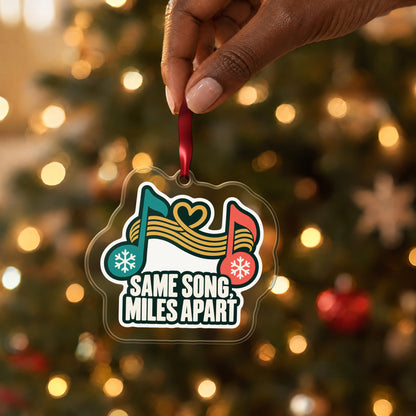 Same Song, Miles Apart Long Distance clear acrylic Christmas ornament held by its red ribbon in a hand, shown at its real size