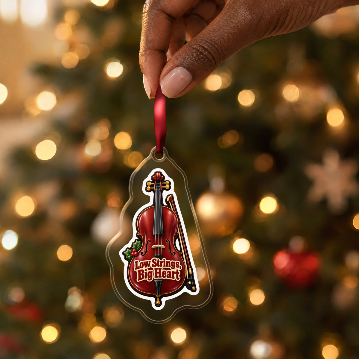 Low Strings, Big Heart Cello clear acrylic Christmas ornament held by its red ribbon in a hand, shown at its real size