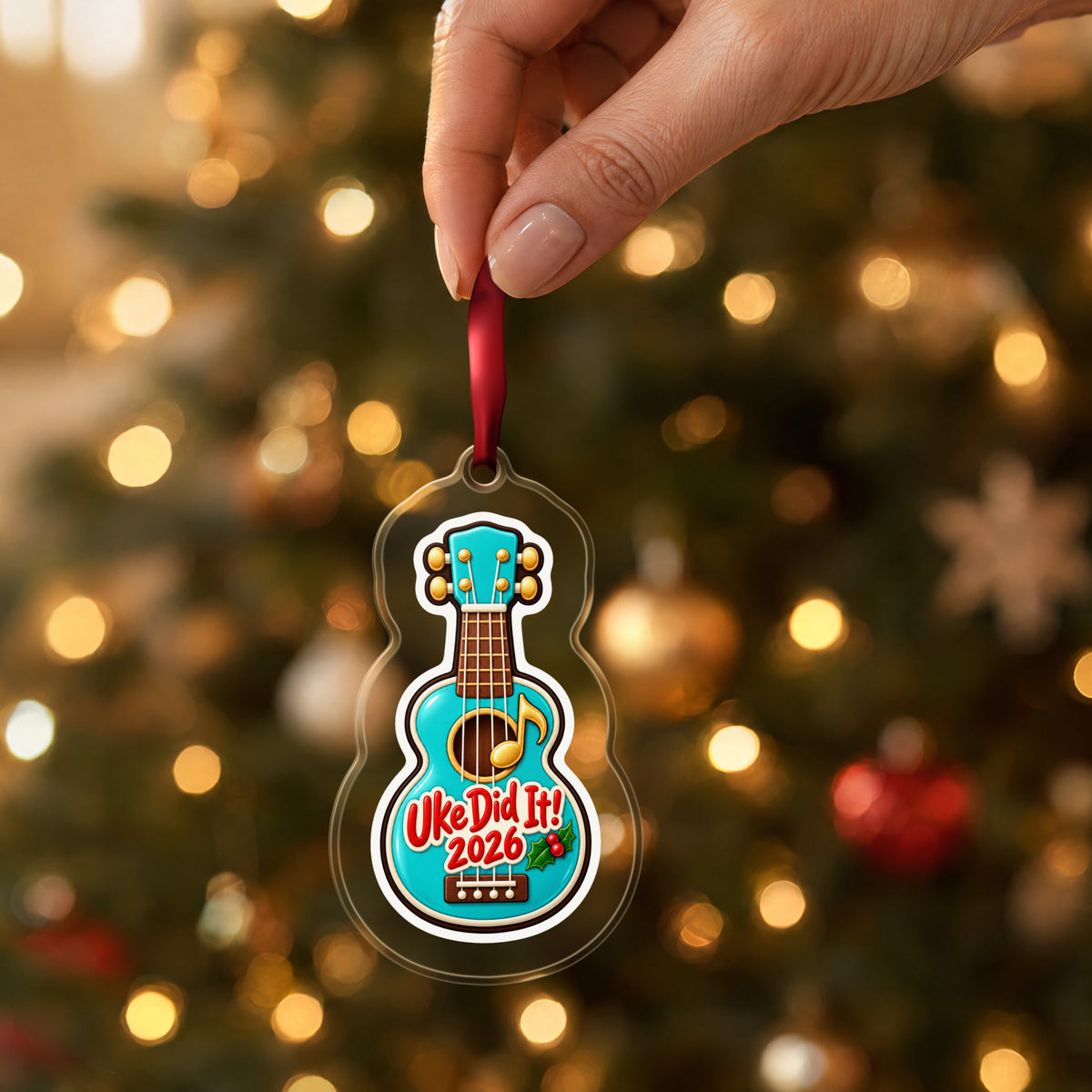 Uke Did It 2026 First Ukulele clear acrylic Christmas ornament held by its red ribbon in a hand, shown at its real size