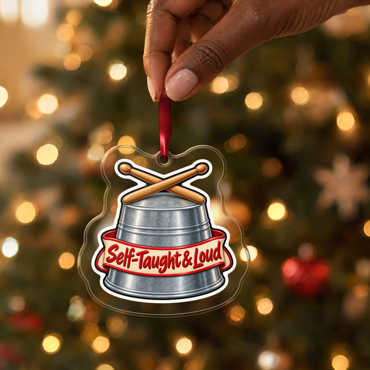 Self-Taught & Loud Bucket Drummer clear acrylic Christmas ornament held by its red ribbon in a hand, shown at its real size
