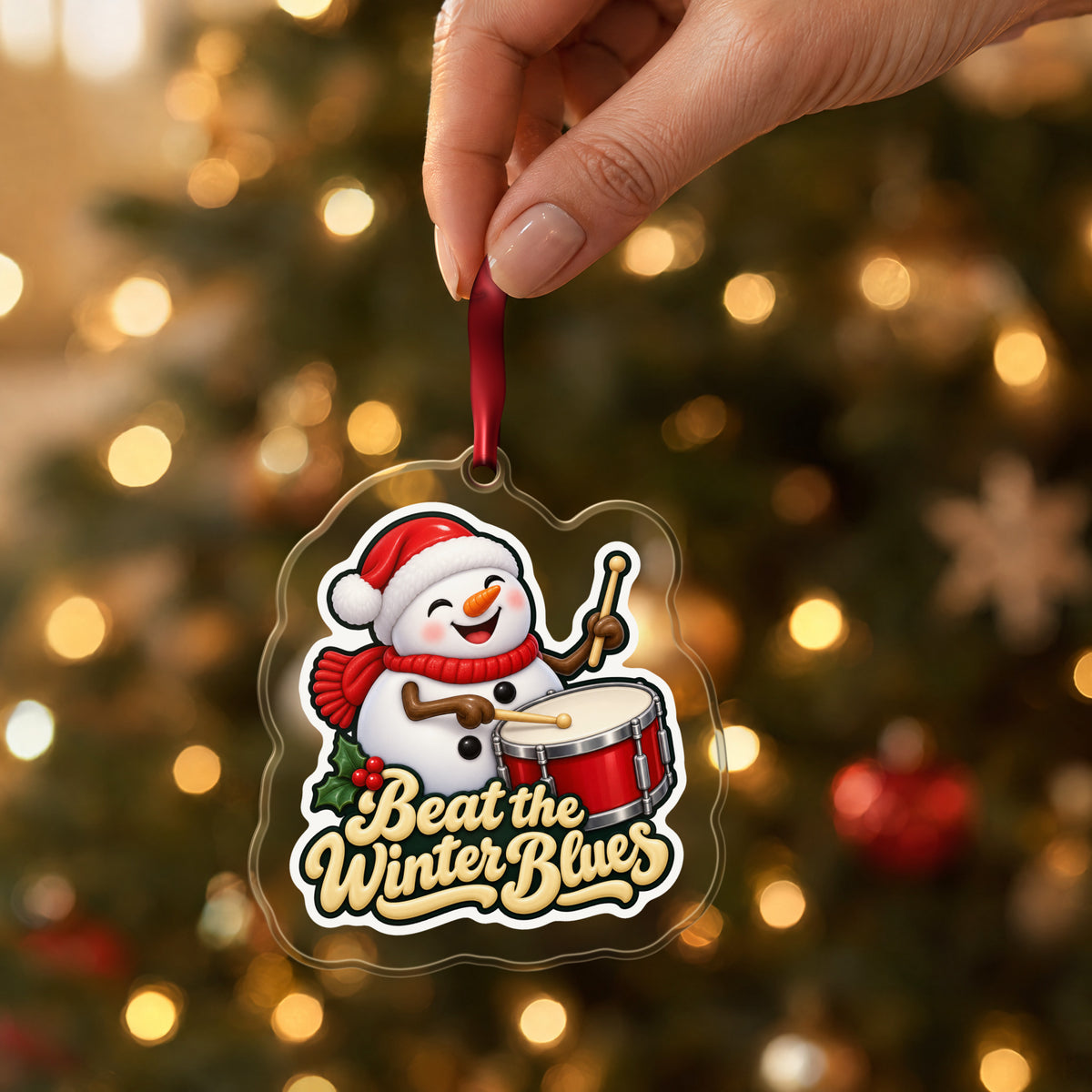 Beat the Winter Blues Snowman Drummer clear acrylic Christmas ornament held by its red ribbon in a hand, shown at its real size