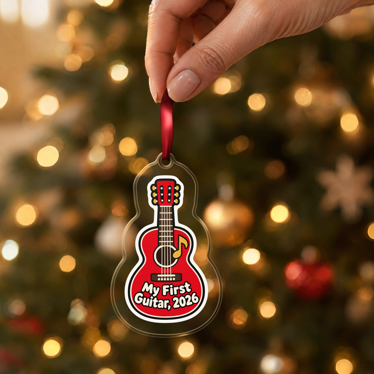 My First Guitar 2026 clear acrylic Christmas ornament held by its red ribbon in a hand, shown at its real size