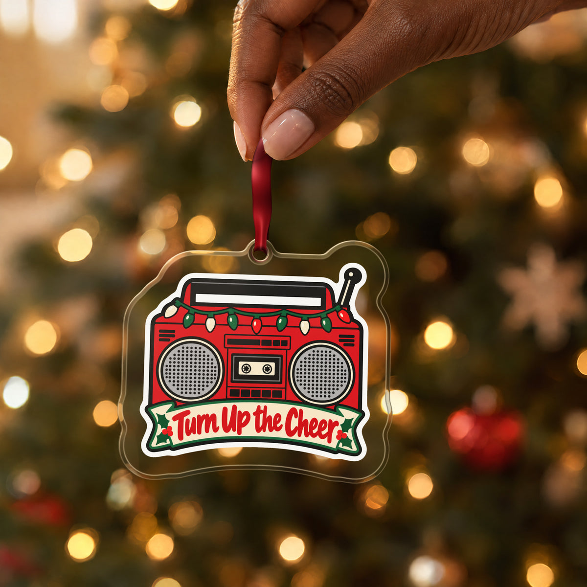 Turn Up the Cheer Boombox clear acrylic Christmas ornament held by its red ribbon in a hand, shown at its real size