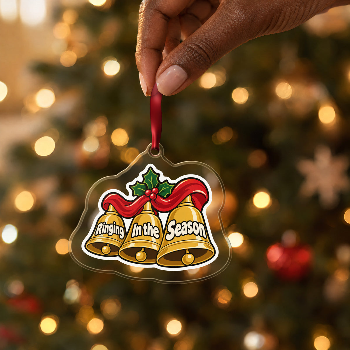 Ringing In the Season Handbells clear acrylic Christmas ornament held by its red ribbon in a hand, shown at its real size