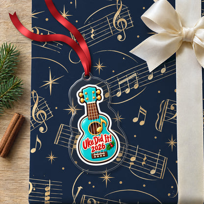 Uke Did It 2026 First Ukulele clear acrylic Christmas ornament lying on a gift wrapped in Rhythm and Beat music wrapping paper