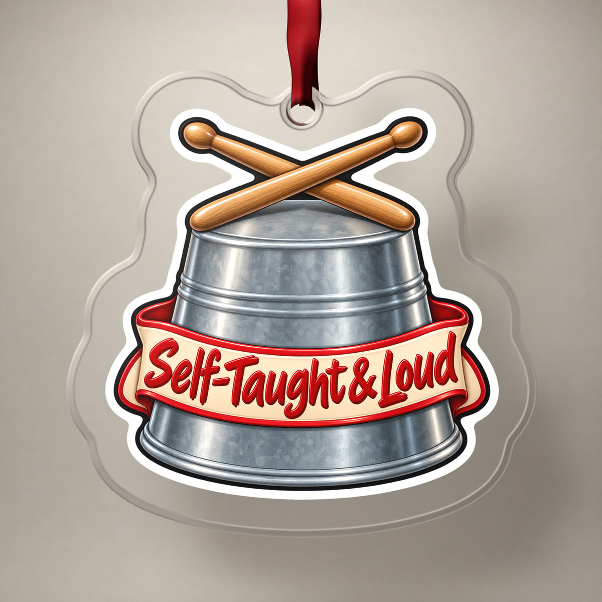 Self-Taught & Loud Bucket Drummer clear acrylic Christmas ornament on a plain studio backdrop with its red ribbon