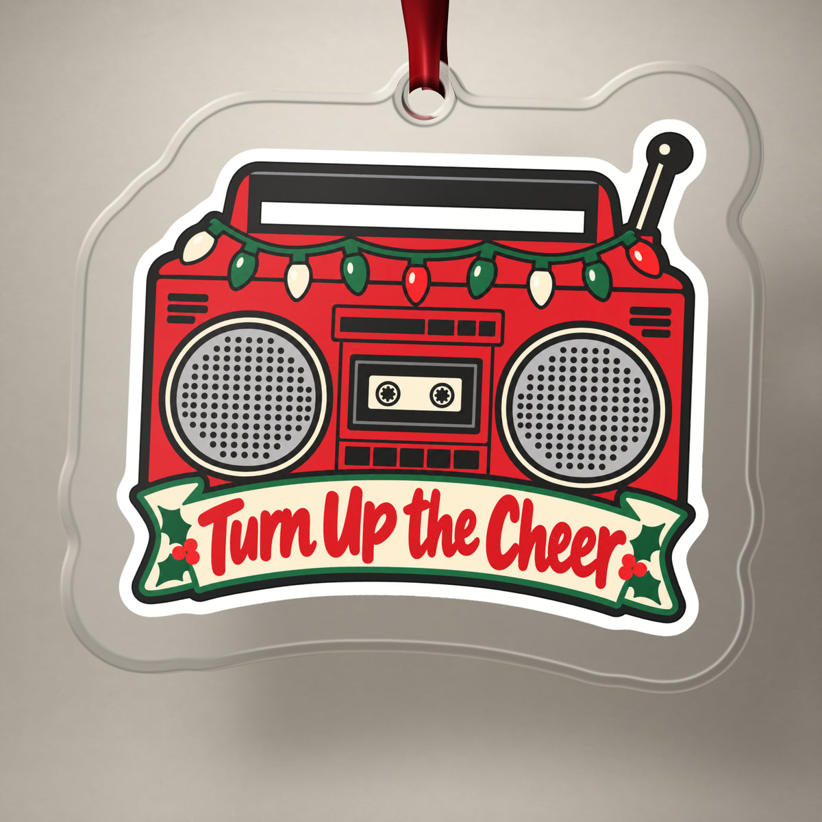 Turn Up the Cheer Boombox clear acrylic Christmas ornament on a plain studio backdrop with its red ribbon