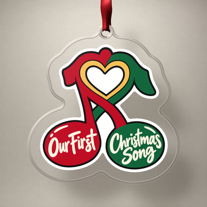 Our First Christmas Song Music Notes clear acrylic Christmas ornament on a plain studio backdrop with its red ribbon
