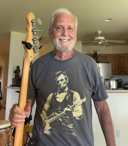 Stephen K. smiling in the grey Lincoln Guitar tee, holding a bass guitar
